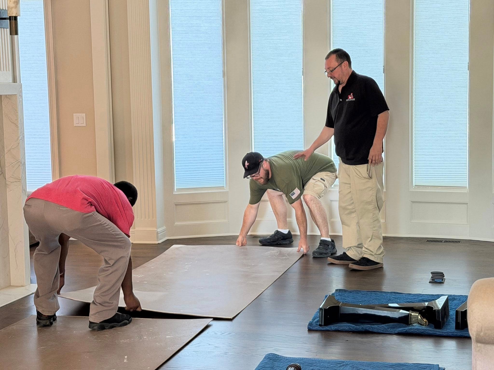 Three men placing large protective sheets on a dark wood floor in a sunlit room.