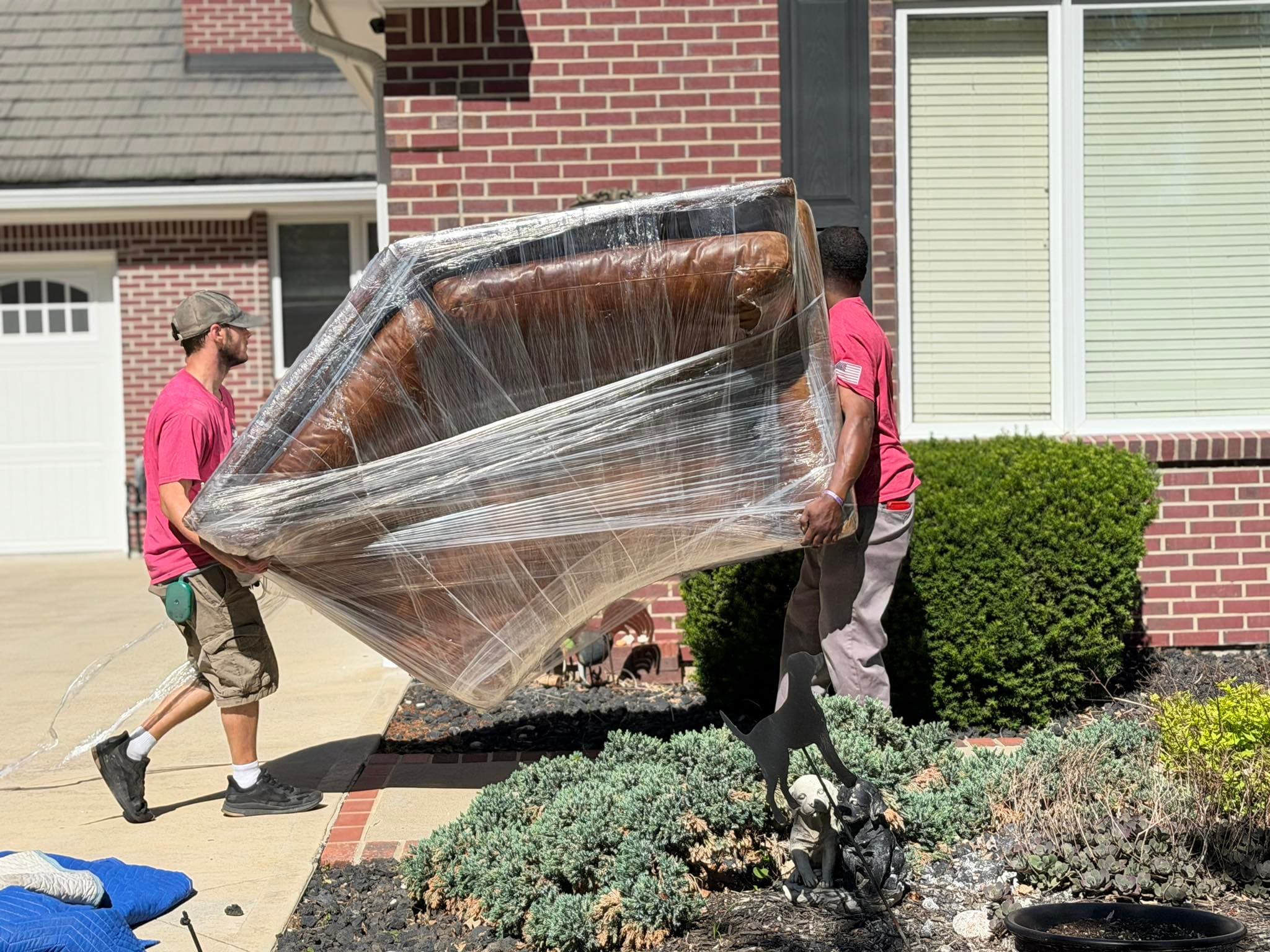 Two movers in red shirts carry a plastic-wrapped leather armchair outside a brick home.