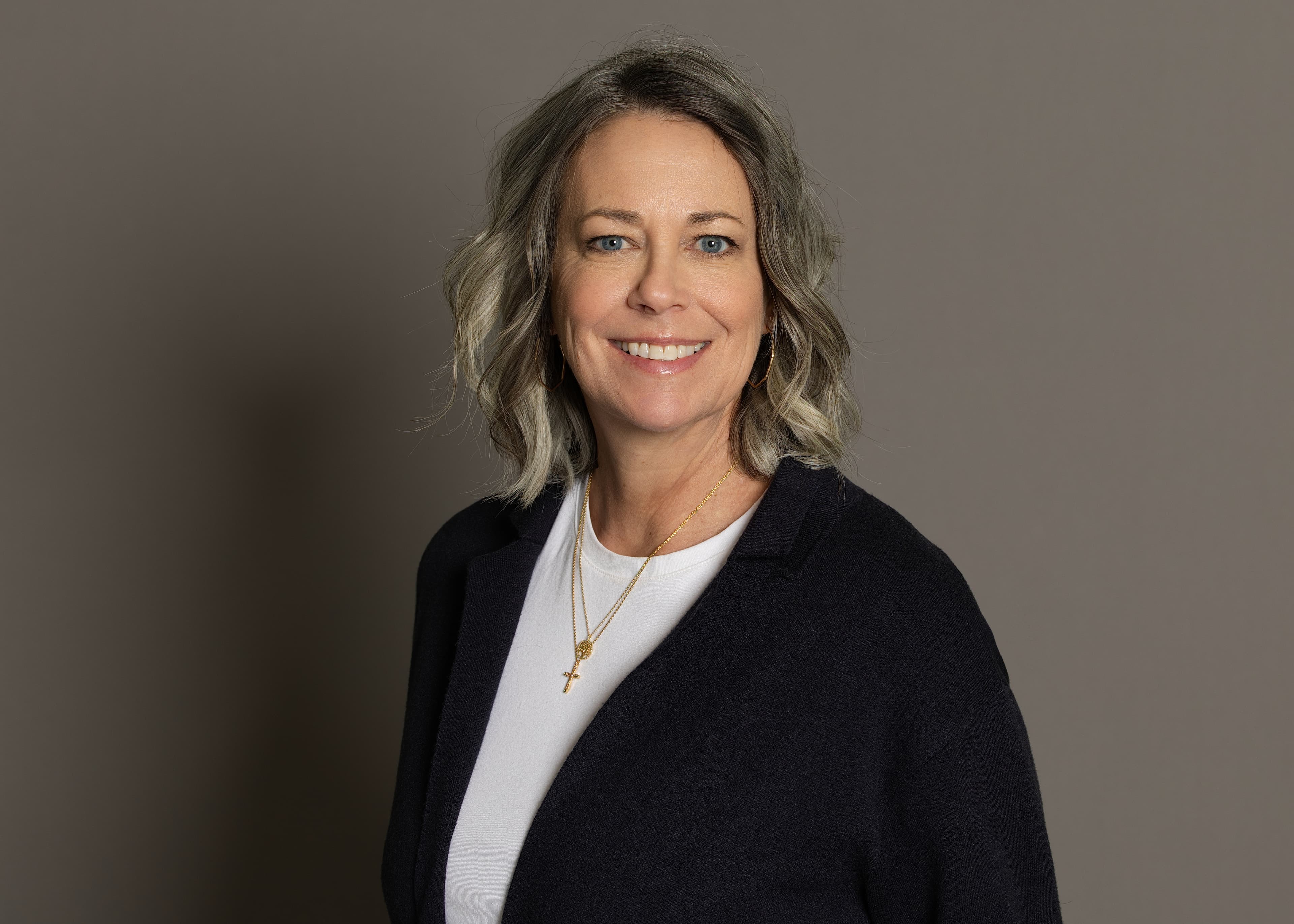 Smiling middle-aged woman with wavy grey hair wearing a navy cardigan and gold necklace.