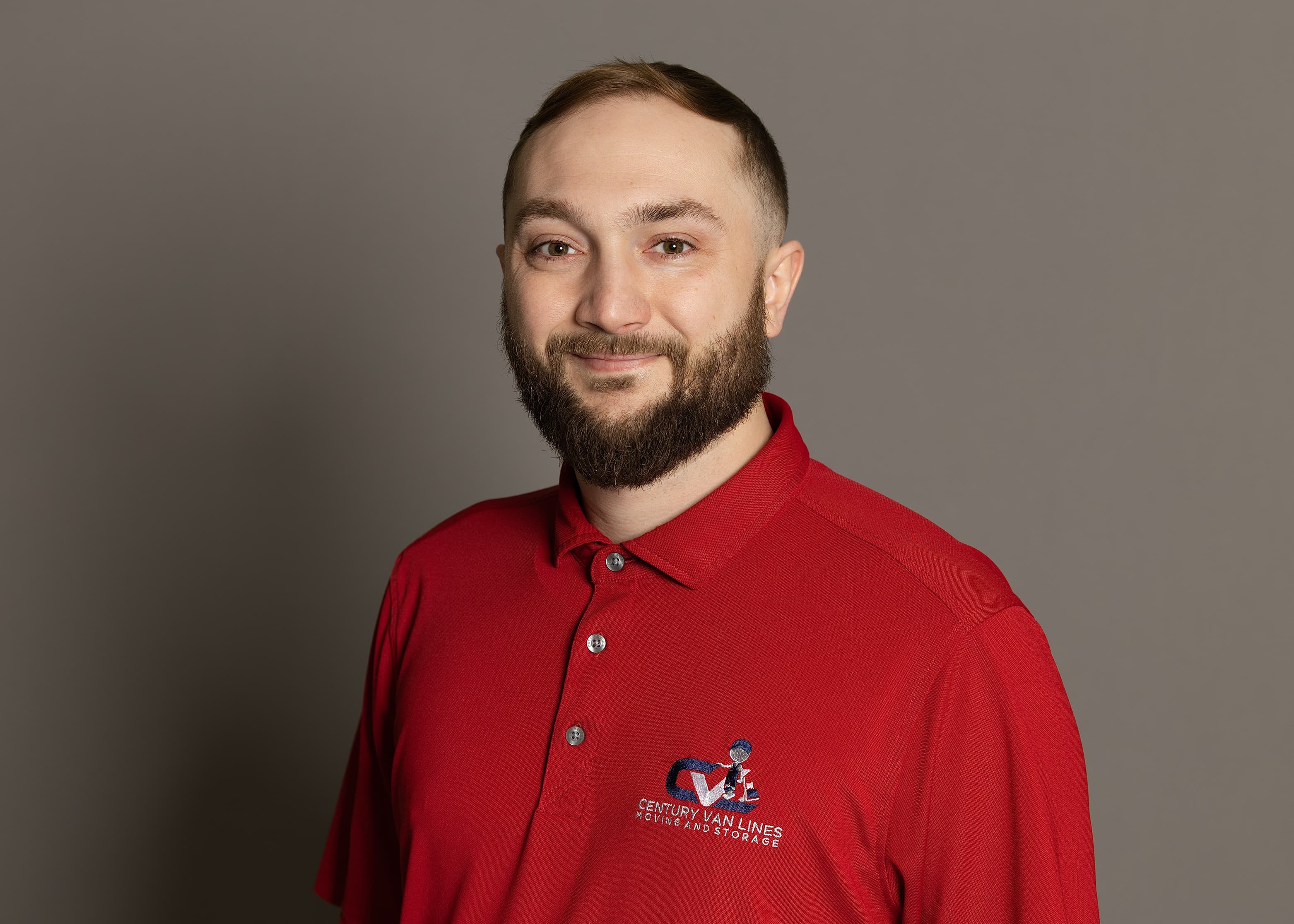 Smiling bearded man in a red Century Van Lines polo shirt against a grey background.