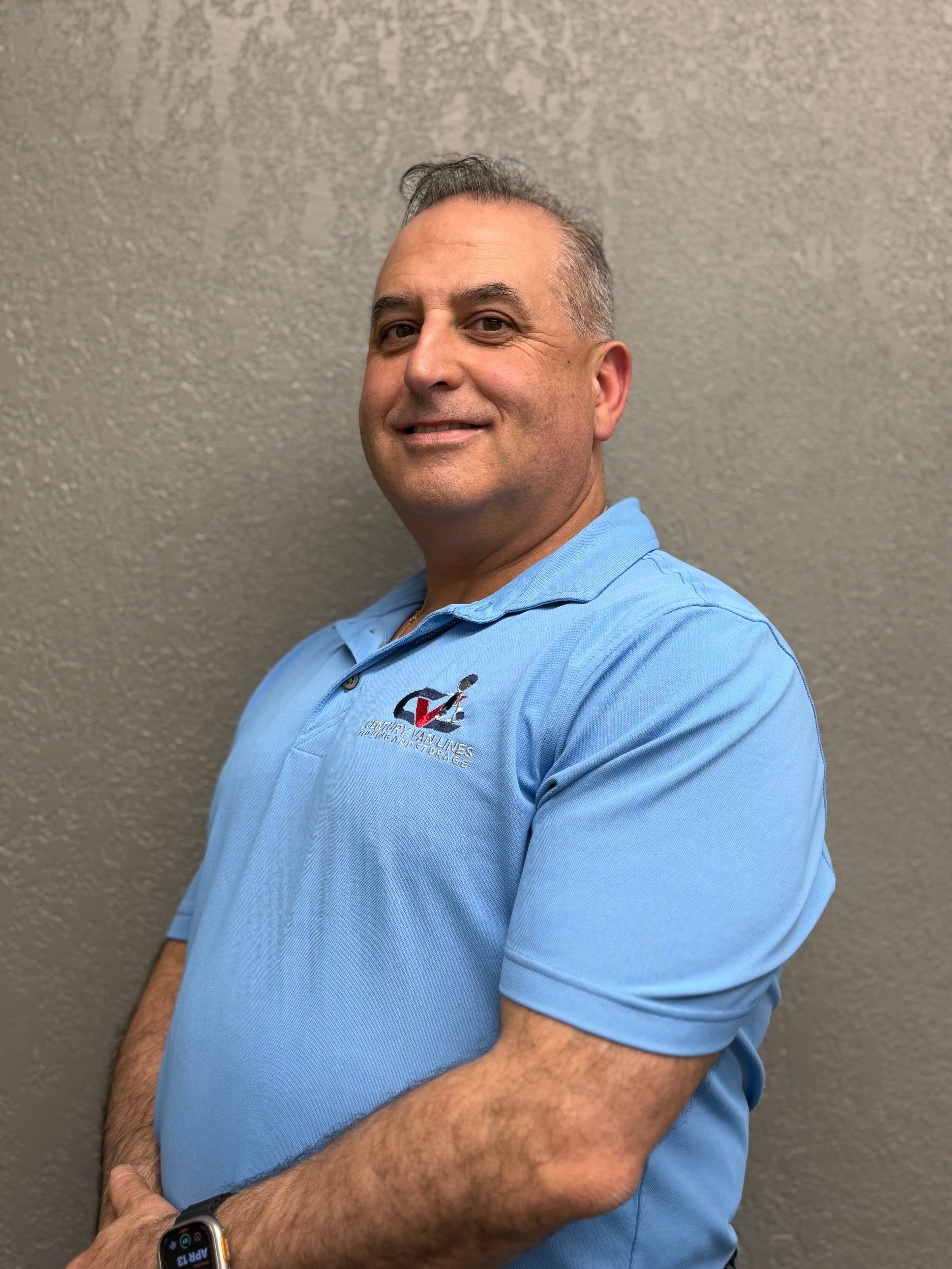 Smiling man in a light blue polo shirt with a company logo against a wall.