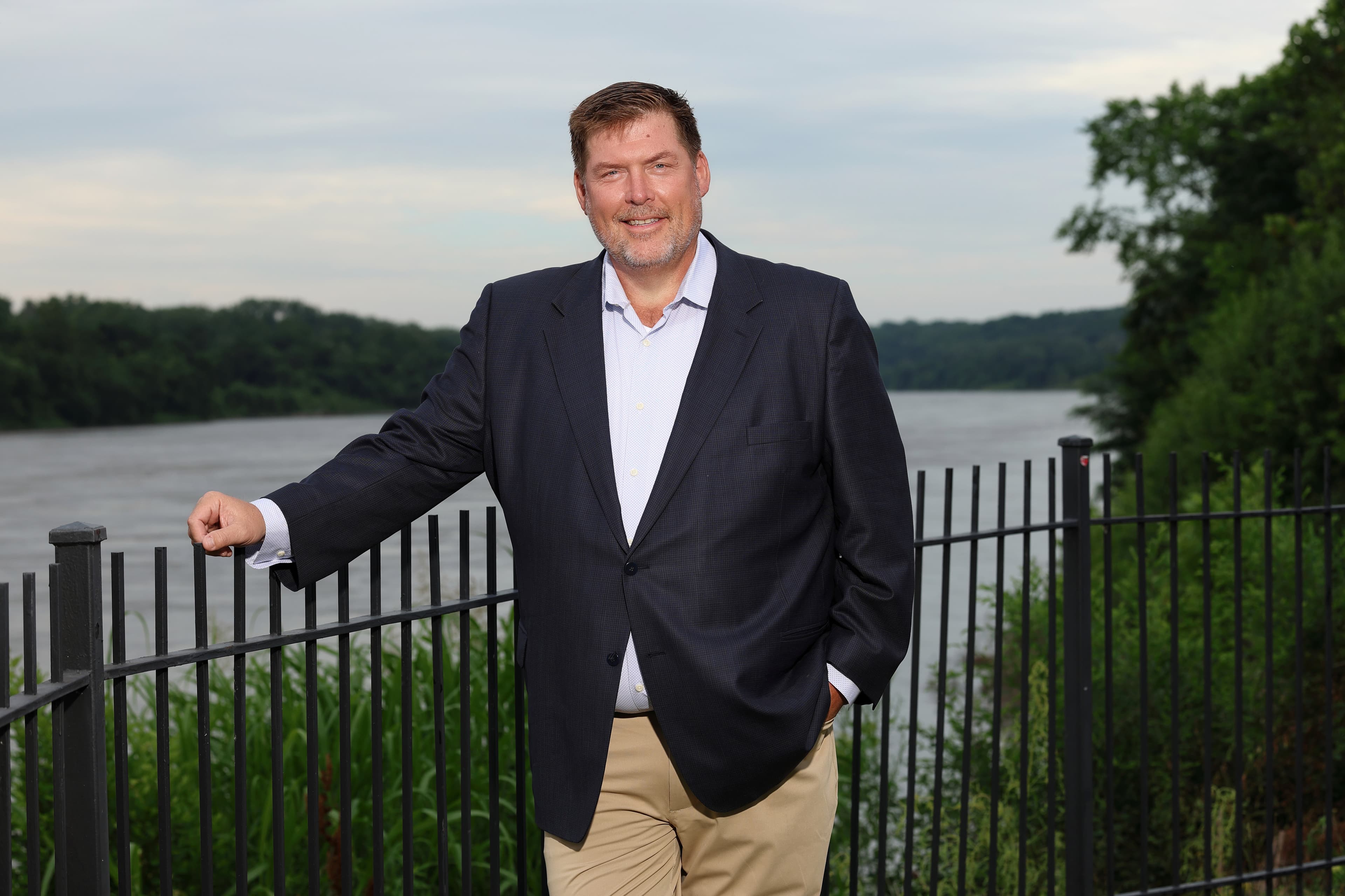 Smiling man in a navy blazer and khakis leaning on a fence overlooking a river.