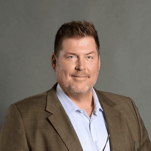 Professional headshot of a smiling man with brown hair and beard wearing a brown blazer.