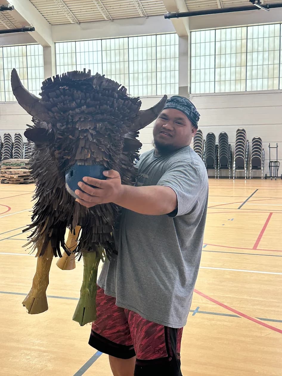 Smiling man in a bandana holds a large fringed bison sculpture in a gymnasium.